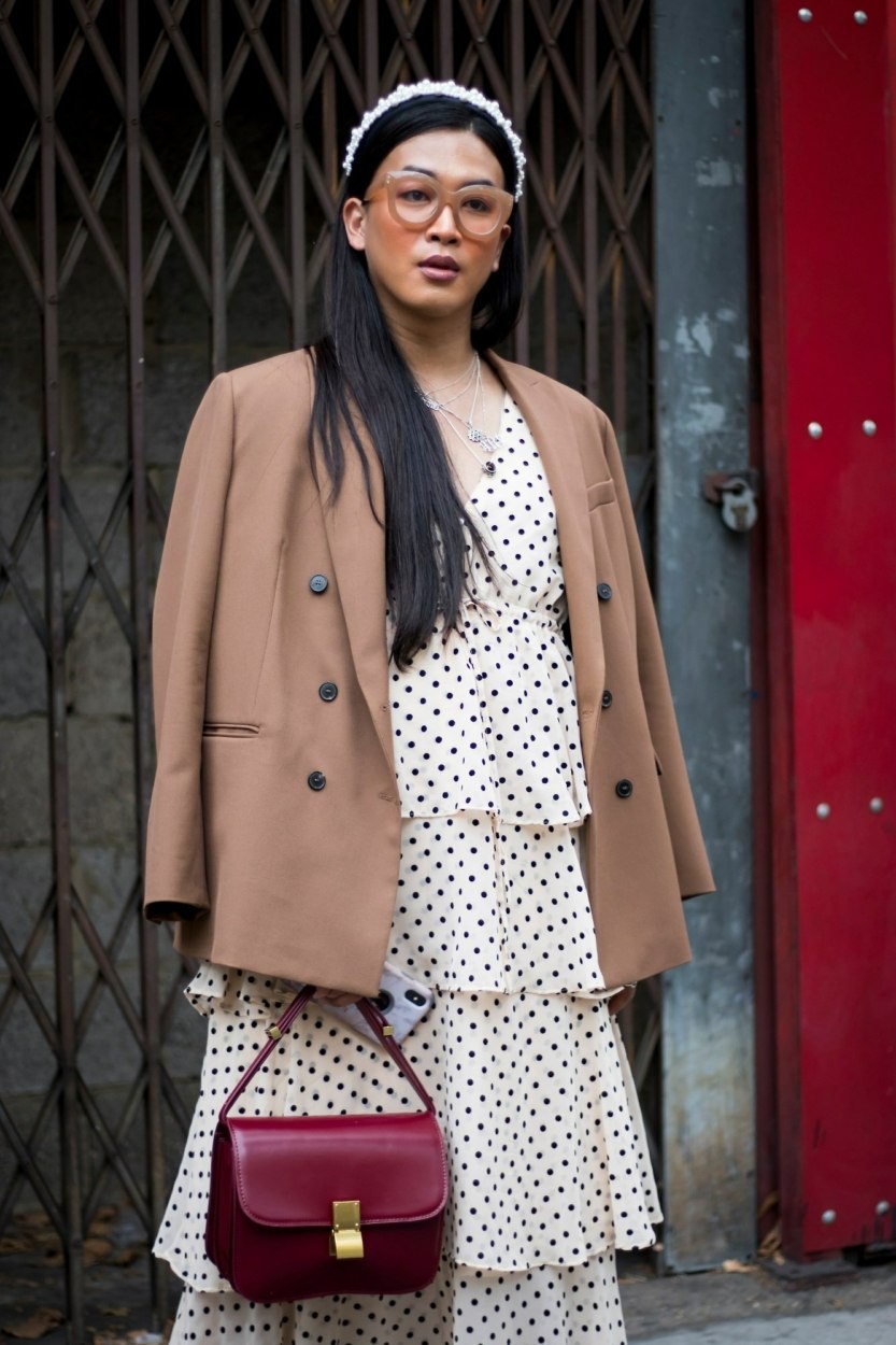 Street style de Fashion Week à Londres – Femme portant une robe étagée à pois noirs sur fond blanc, sous une veste marron clair ou beige foncé, portant un sac rouge cerise